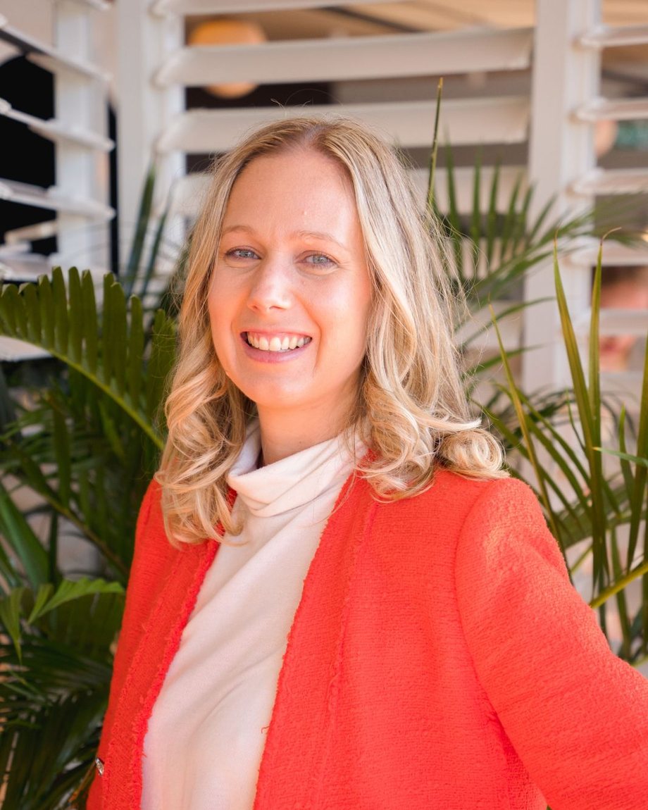 Angie Farmer standing in front of an indoor palm, smiling, in a coral blazer and cream top