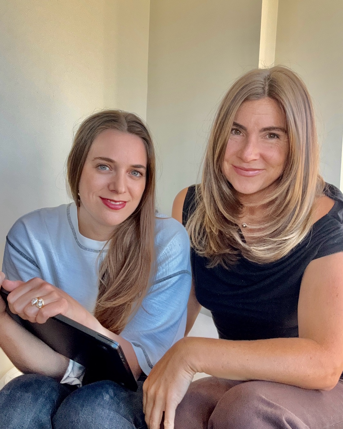 Jami Emerson Cheneval and Cortney Thomson seated side by side against a pale wall, one holding a tablet